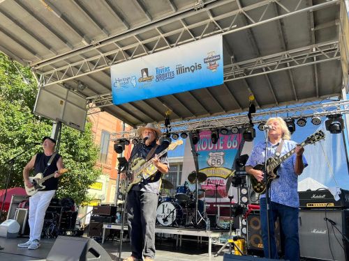 A band performs on stage at a festival, with musicians playing guitar and singing under a sunny sky.