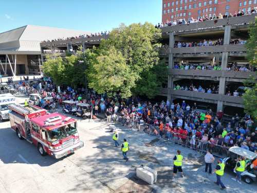A large crowd gathers along a street for a parade, with a fire truck and people in safety vests visible in the foreground.