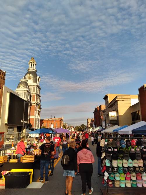 A bustling street market with vendors, shoppers, and a historic building under a blue sky with scattered clouds.