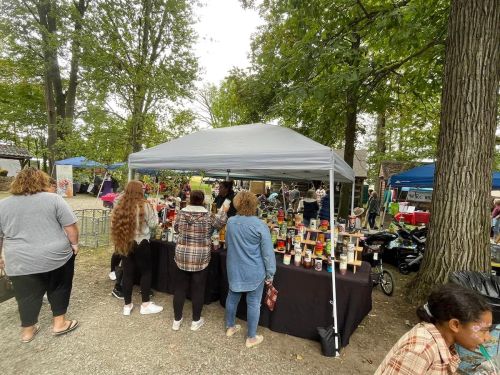 A group of people browsing a market stall with various candles, surrounded by trees and other vendor tents.