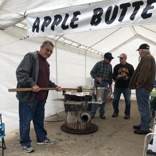 A man stirs a pot at an outdoor event under a sign that reads "APPLE BUTTER," with others chatting nearby.