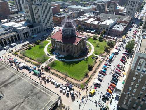 Aerial view of a bustling city square with a historic building, green lawn, and cars lined up for an event.