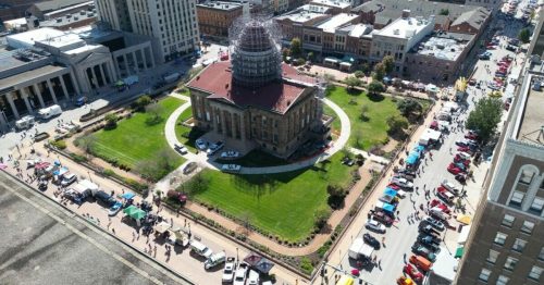 Aerial view of a bustling city square with a historic building, green lawn, and crowds at a community event.