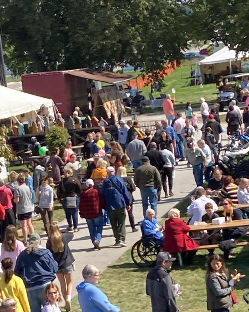 A bustling outdoor festival scene with people walking, chatting, and enjoying activities among tents and food stalls.