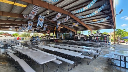 A spacious outdoor pavilion with long tables, decorated with banners, and surrounded by food stalls and palm trees.