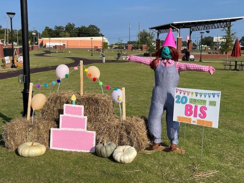 A scarecrow in a party hat stands by hay bales and a festive sign, celebrating a birthday with colorful decorations.