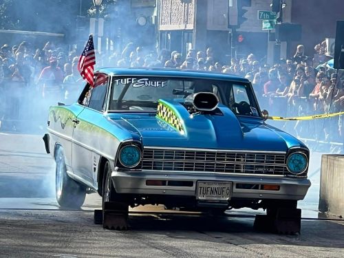 A blue classic car with a hood scoop and American flag drives through smoke, surrounded by a cheering crowd.