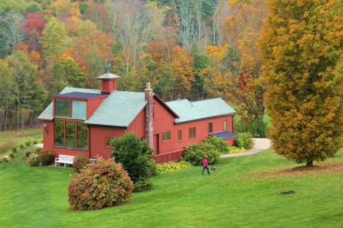 A red house surrounded by colorful autumn trees, with a person walking a dog on a path nearby.