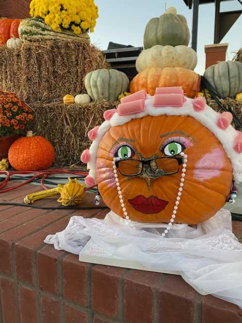 A decorated pumpkin with glasses, lipstick, and pearls, sitting among colorful pumpkins and hay bales.