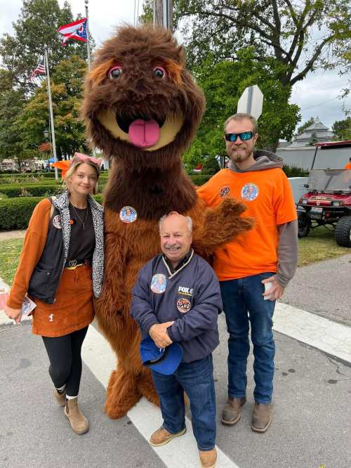 A furry mascot poses with two people in orange shirts at an outdoor event, surrounded by trees and flags.