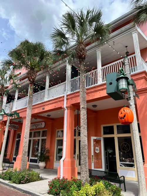 Colorful storefronts with palm trees and festive decorations, including a pumpkin, under a cloudy sky.