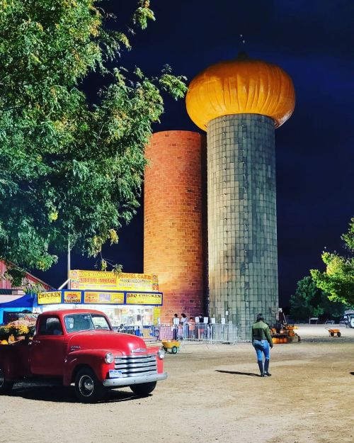A vintage red truck parked near large silos under a night sky, with colorful lights and a festive atmosphere.