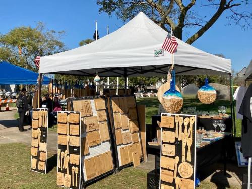 A craft booth with wooden items displayed under a white tent, featuring small American flags and a sunny outdoor setting.