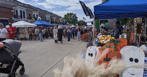 A bustling outdoor market with vendors, colorful tents, and autumn decorations, filled with people enjoying the day.