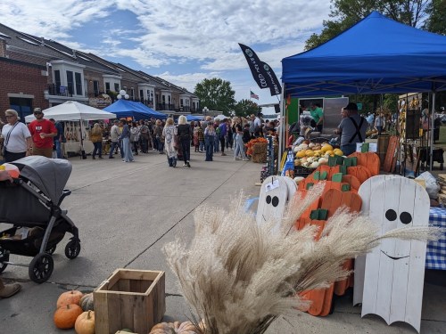 A bustling outdoor market with vendors, colorful tents, pumpkins, and a crowd enjoying the festivities on a sunny day.