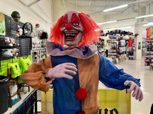 A creepy clown mannequin with red hair and a sinister grin, displayed in a store filled with Halloween decorations.