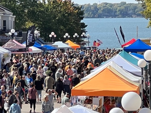 A bustling outdoor market with colorful tents and a large crowd by a lake on a sunny day.