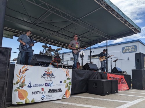 A band performs on stage at a festival, with a banner displaying sponsors in front of them.