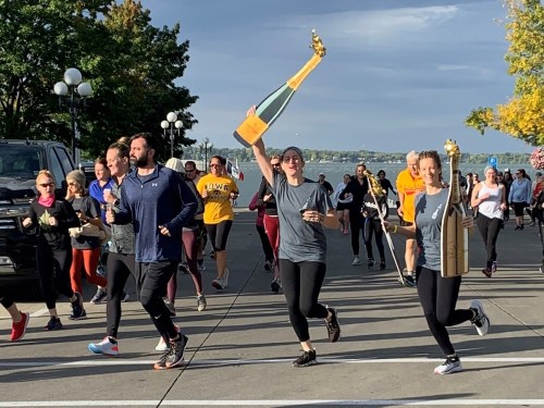 A group of runners celebrates with oversized champagne bottles during a fun run by a waterfront.