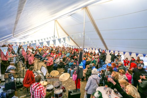 A lively crowd enjoys a performance under a large tent, with musicians in plaid shirts and festive decorations.