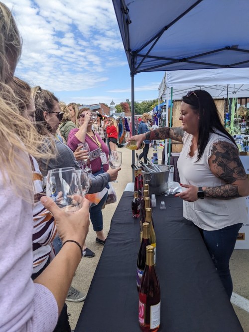 A vendor pours wine for customers at an outdoor market, with people holding glasses and enjoying the event.