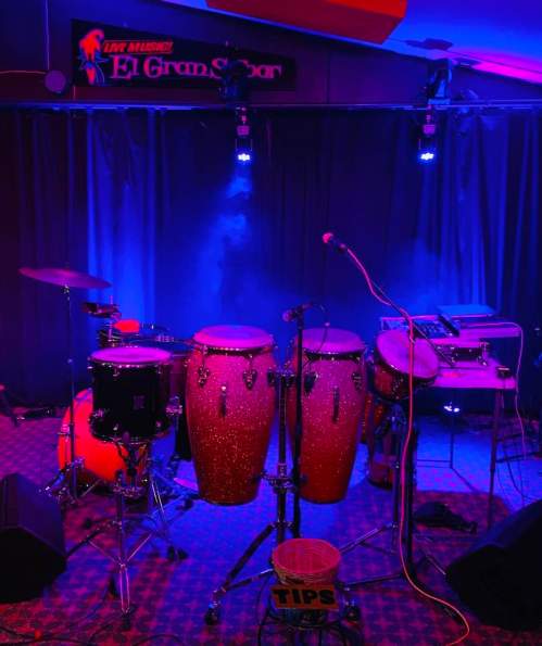 A stage setup with drums, congas, and a microphone, illuminated by blue lights in a music venue.