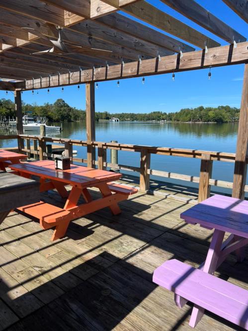 A wooden deck with colorful picnic tables overlooks a calm lake surrounded by trees under a clear blue sky.