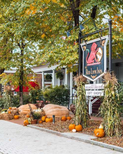 A charming inn sign surrounded by autumn decorations, including pumpkins and hay bales, under colorful trees.