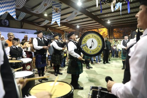 A band in traditional attire performs with bagpipes and drums at a lively indoor event, surrounded by an audience.