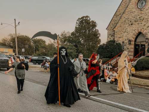 A Halloween parade featuring costumed characters, including a grim reaper, walking along a street near a stone church.
