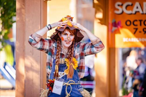 A person dressed as a scarecrow, with braids and autumn decorations, poses playfully at an outdoor event.