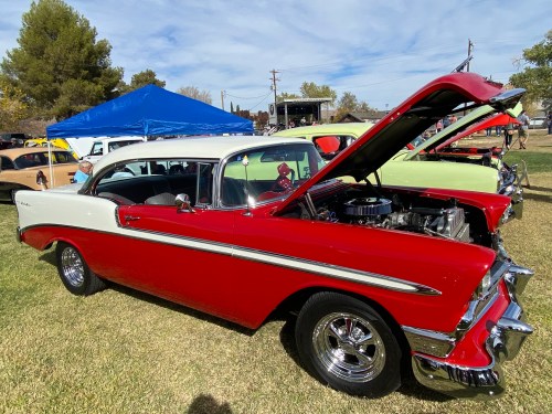 A classic red and white 1956 car with the hood open, parked on grass at a car show with other vehicles in the background.