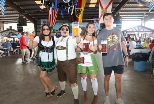 Four people in festive attire hold up large beer mugs at a lively indoor event decorated with flags and banners.