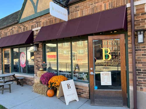 Exterior of a botanical bar with a sign, large windows, and fall decorations including pumpkins and mums.