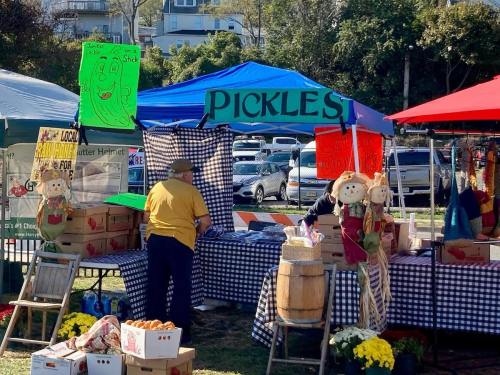A vendor at a market booth labeled "PICKLES" with colorful signs and decorations, surrounded by boxes and scarecrows.