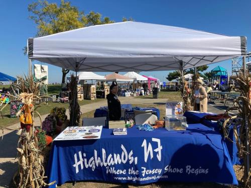 A vendor booth at an outdoor event with a blue tablecloth displaying "Highlands, NJ" and decorations made of corn stalks.
