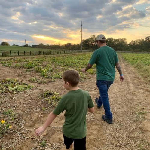 A man and a boy walk through a field, surrounded by rows of plants under a cloudy sky.