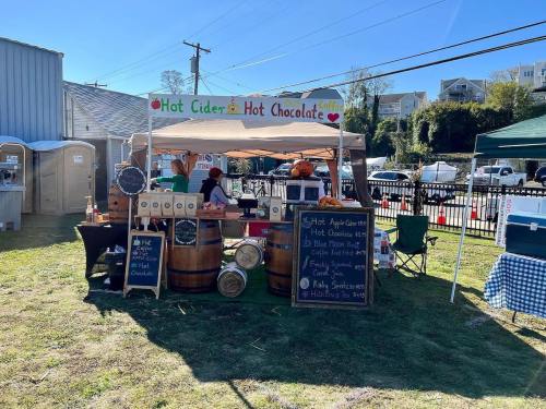 A cozy outdoor stand selling hot cider and hot chocolate, with barrels and a chalkboard menu.
