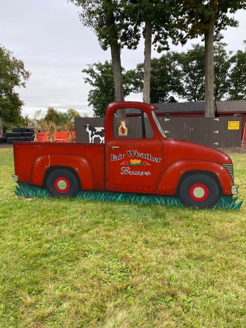 A vintage red pickup truck cutout with "Fair Weather" written on it, set on green grass in a park-like area.