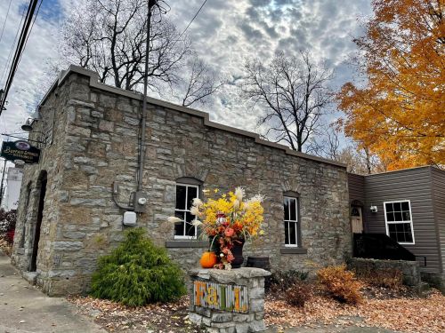 A stone building with autumn decorations, surrounded by trees and a cloudy sky. Fall leaves cover the ground.