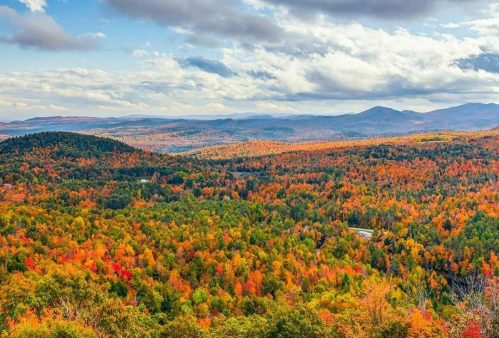 A vibrant autumn landscape with colorful trees, rolling hills, and a blue sky dotted with clouds.