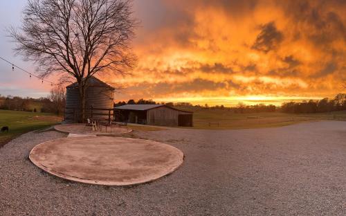 A vibrant sunset casts orange and yellow hues over a rural landscape with a barn and a tree.