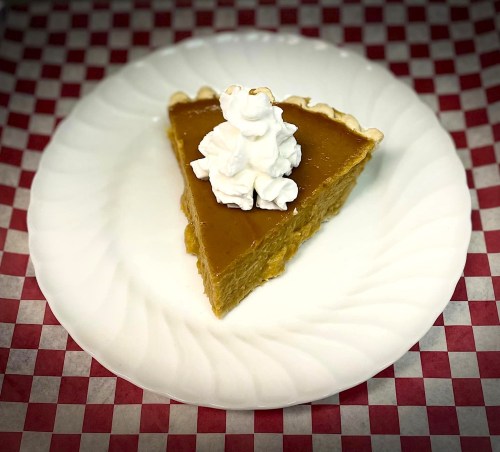 A slice of pumpkin pie topped with whipped cream on a white plate, set against a red and white checkered background.