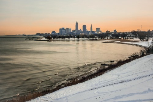 A winter sunset over a snowy shoreline, with the Cleveland skyline in the background and calm waters in the foreground.
