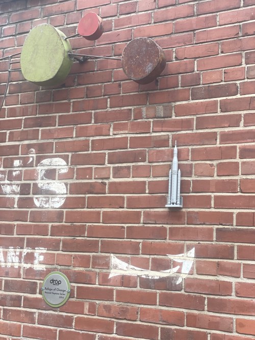 Colorful circular shapes and a silver sculpture are mounted on a brick wall, with a sign below.