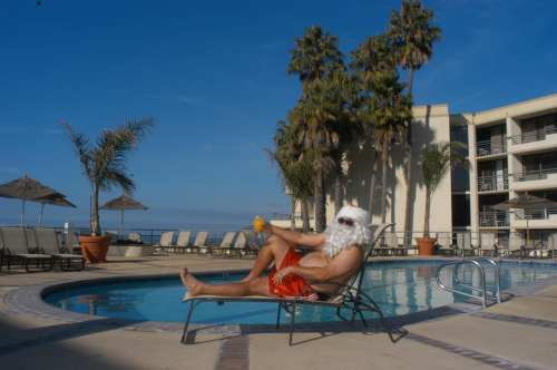 A man in a Santa costume relaxes by a pool, sipping a drink, with palm trees and a sunny sky in the background.