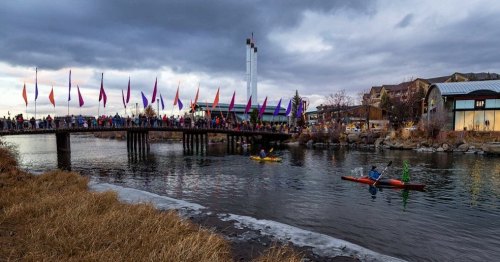 A lively scene by a river with a bridge, colorful flags, and kayakers, set against a cloudy sky.