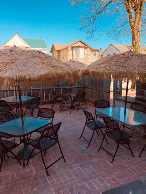 Outdoor seating area with straw umbrellas and tables on a brick patio, surrounded by wooden fencing and houses.