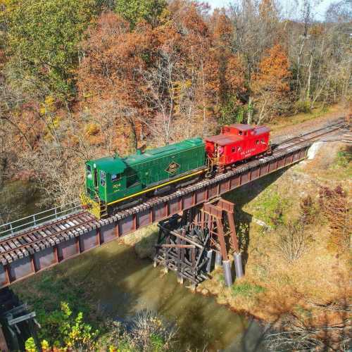 A green locomotive and red caboose cross a bridge over a creek, surrounded by autumn foliage.