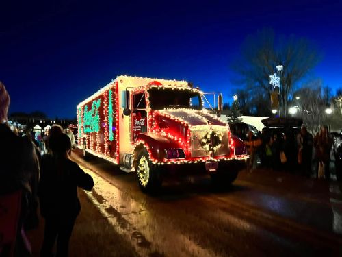 A brightly lit holiday truck decorated with lights and festive designs drives past a crowd at dusk.
